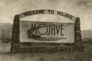 Sepia-toned black-and-white photograph of an early “Welcome to Mojave” wooden sign framed by stone pillars. The sign stands in the open desert with wagon tracks in the foreground and distant hills behind. The image features fine film grain, soft light, and faded edges, evoking the authentic look of an 1800s glass-plate photograph from the early Mojave settlement era.